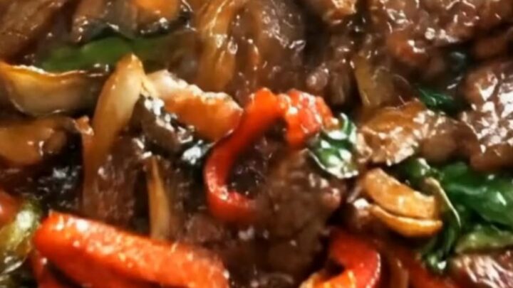 A close-up shot of a saucy pepper steak stir-fry in a black skillet with vibrant red peppers and onions.