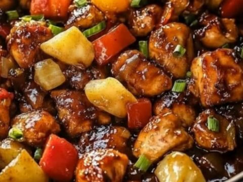 A glossy, sweet and sticky pineapple chicken skillet in a black pan with vibrant red peppers.