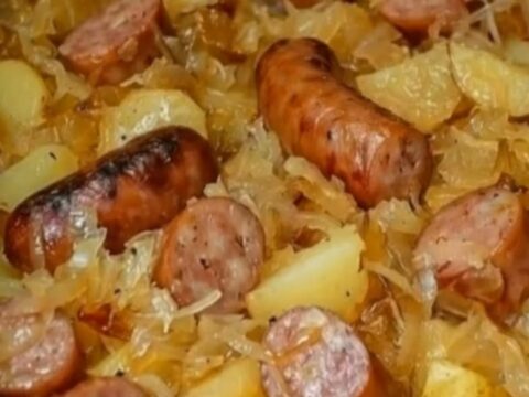 A steaming skillet filled with sliced kielbasa, golden potato chunks, and sauerkraut on a wooden table.