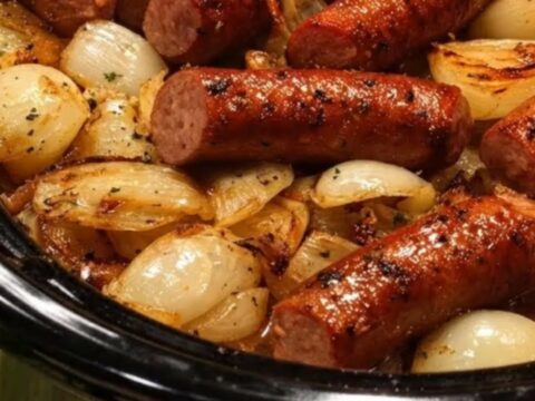 A close-up of juicy, browned sausage links and golden caramelized onions in a black slow cooker.