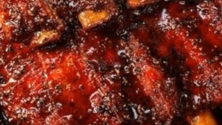 A close-up of sticky, caramelized BBQ ribs resting in a foil-lined baking pan, showing a gleaming sauce.