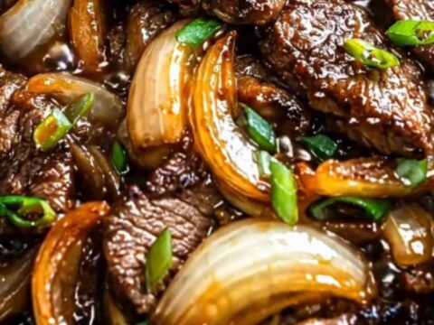 A close-up of glossy beef slices and caramelized onions in a cast-iron skillet, with steam rising.