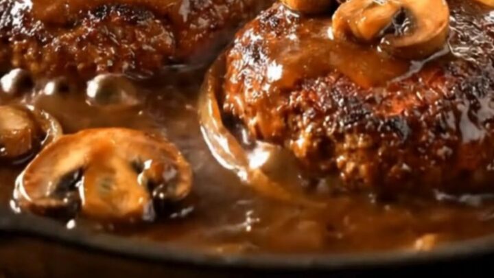 A cast-iron skillet filled with juicy hamburger steaks covered in a rich, dark mushroom gravy.