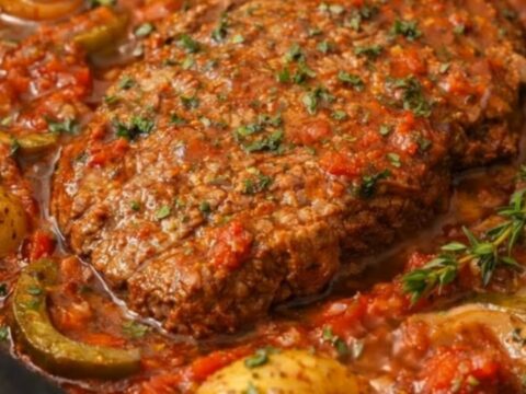 Tender Swiss steak coated in a rich tomato, onion, and bell pepper sauce served on a white plate.