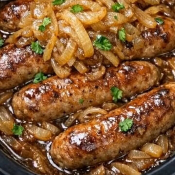 A close-up of juicy pork sausages and caramelized onions in a rich brown gravy served with creamy mashed potatoes.