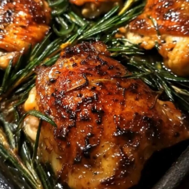 A golden-brown roasted chicken thigh with crispy skin, garnished with fresh rosemary on a dark baking pan.