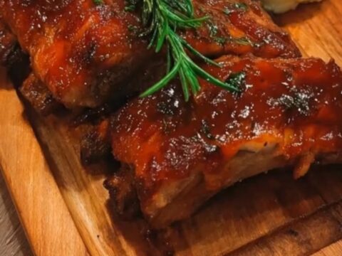 A close-up of glossy, caramelized sticky honey garlic ribs next to a heap of creamy mashed potatoes on a wooden board.
