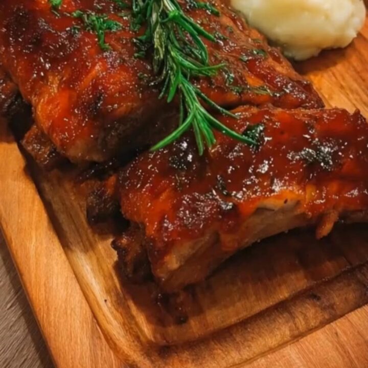 A close-up of glossy, caramelized sticky honey garlic ribs next to a heap of creamy mashed potatoes on a wooden board.