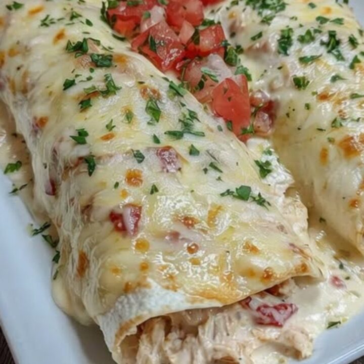 A white rectangular platter holding two golden, cheese-blanketed chicken enchiladas topped with fresh diced tomatoes