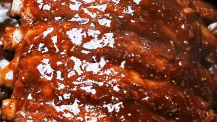 A close-up of tender barbecue ribs being lifted from a black slow cooker with metal tongs.