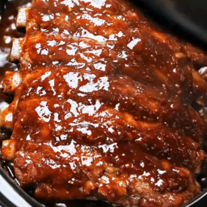 A close-up of tender barbecue ribs being lifted from a black slow cooker with metal tongs.