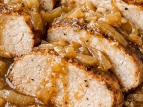 A close-up of sliced pork tenderloin smothered in a rich, glossy caramelized onion gravy in a white baking dish.