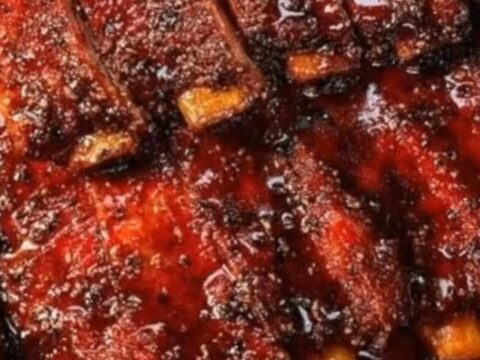 A close-up of deeply caramelized, glistening BBQ ribs resting on a butcher paper-lined tray with rising steam.