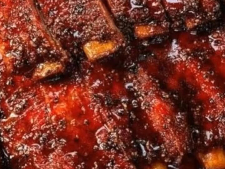 A close-up of deeply caramelized, glistening BBQ ribs resting on a butcher paper-lined tray with rising steam.