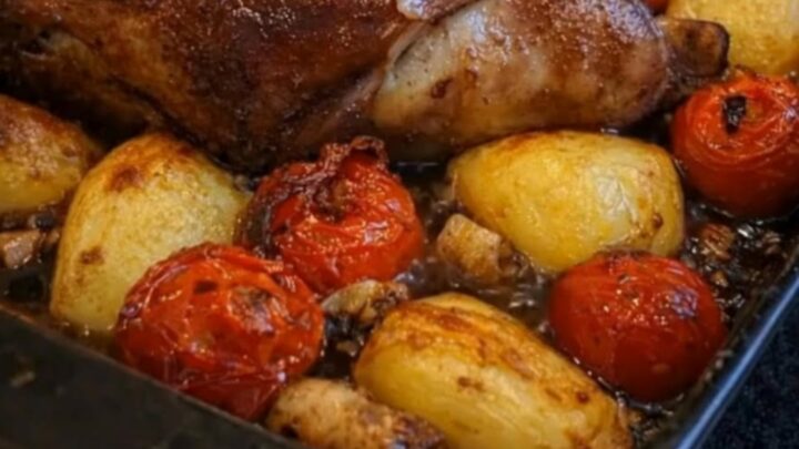 A rustic roasting pan filled with herb-crusted lamb, golden roasted potatoes, and burst cherry tomatoes.