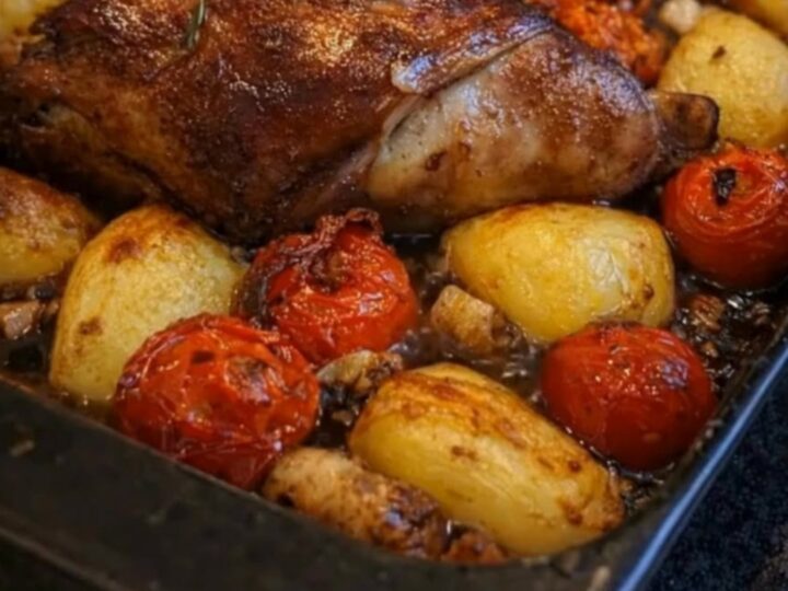 A rustic roasting pan filled with herb-crusted lamb, golden roasted potatoes, and burst cherry tomatoes.