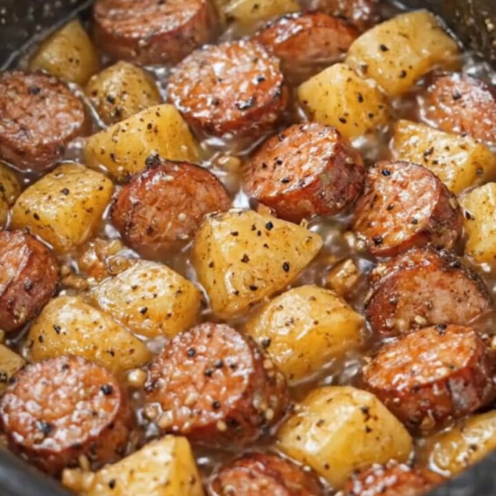 A close-up of a serving spoon scooping steaming sausage and potato chunks from a black slow cooker.