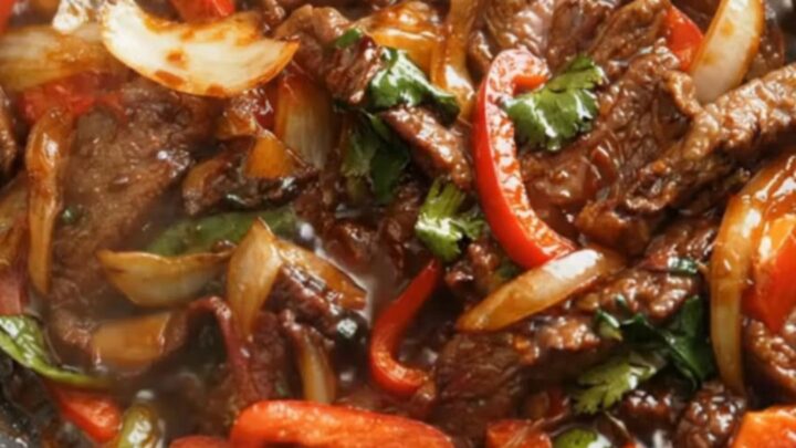 A close-up of glossy, stir-fried beef strips and red bell peppers in a black skillet with steam rising.