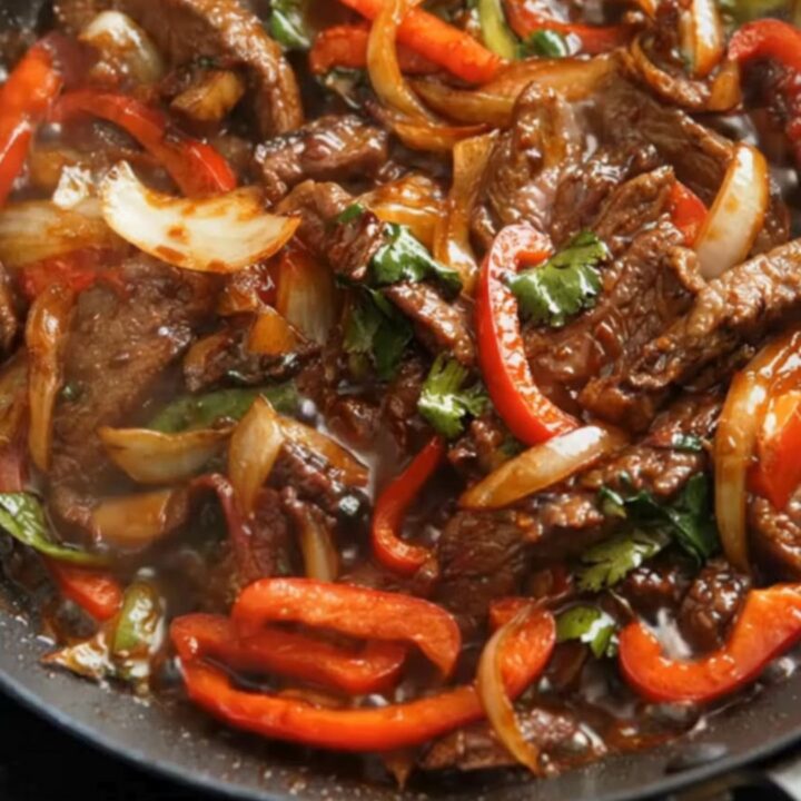 A close-up of glossy, stir-fried beef strips and red bell peppers in a black skillet with steam rising.