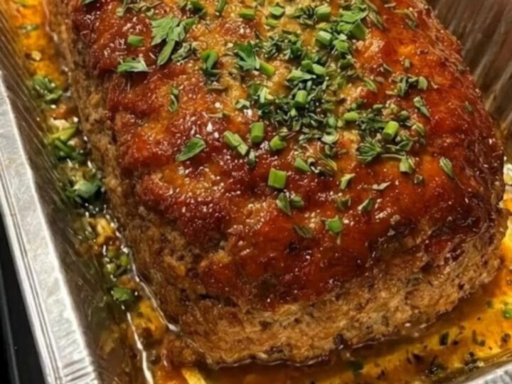 A thick, juicy slice of sweet glazed meatloaf resting on a white plate with fresh green herbs.