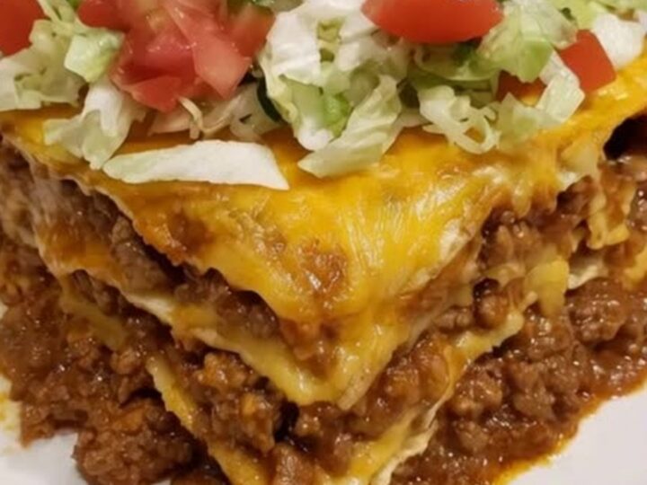 A tall, perfectly layered slice of taco lasagna casserole stack garnished with fresh lettuce, tomatoes, and sour cream.