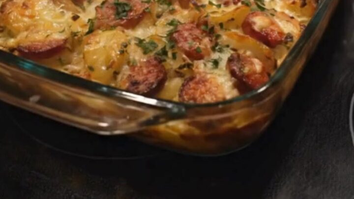 A golden, bubbly cheesy sausage and potato bake in a glass baking dish with a large metal spatula lifting a serving.