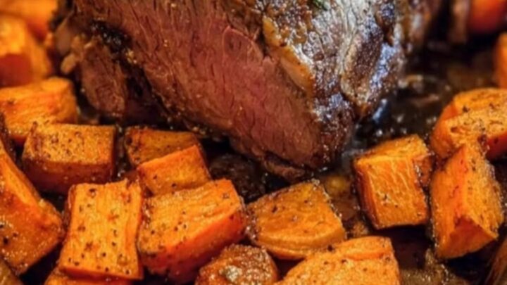 A glistening herb-crusted beef roast sits on a dark baking sheet surrounded by roasted sweet potato cubes.