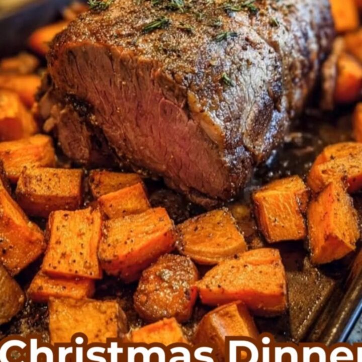 A glistening herb-crusted beef roast sits on a dark baking sheet surrounded by roasted sweet potato cubes.