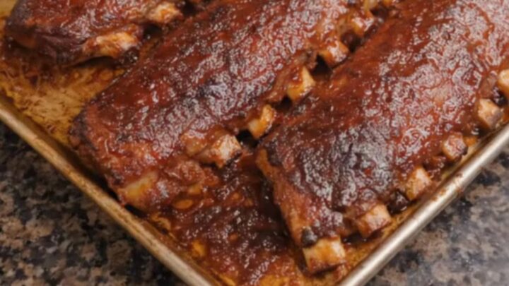 Three glossy, caramelized racks of barbecue ribs sitting on a baking sheet, steaming with juices.