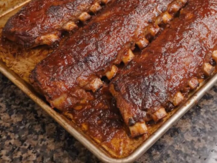 Three glossy, caramelized racks of barbecue ribs sitting on a baking sheet, steaming with juices.