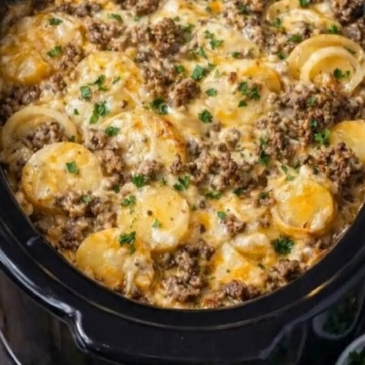 A close-up of a slow cooker filled with layers of tender potatoes, browned beef, and bubbling melted cheese with parsley.