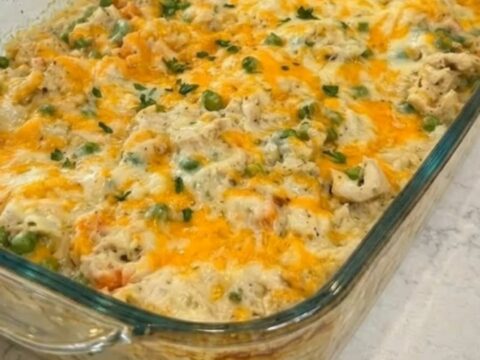 A steaming 9x13 glass baking dish filled with cheesy chicken and rice casserole, topped with melted cheddar and green peas.
