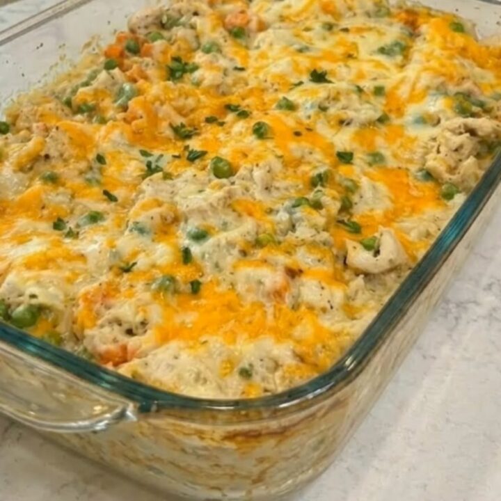 A steaming 9x13 glass baking dish filled with cheesy chicken and rice casserole, topped with melted cheddar and green peas.