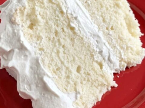 A tall slice of fluffy white layer cake with smooth frosting on a white cake stand.
