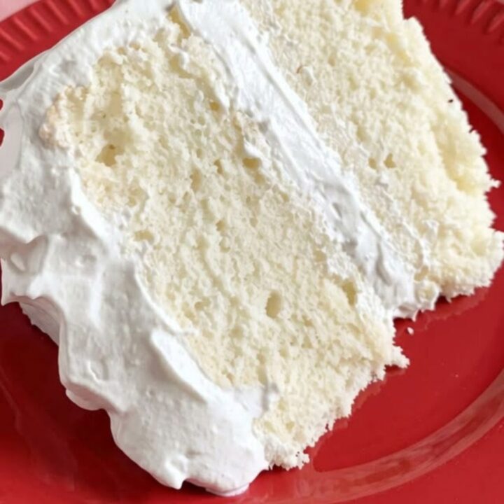A tall slice of fluffy white layer cake with smooth frosting on a white cake stand.