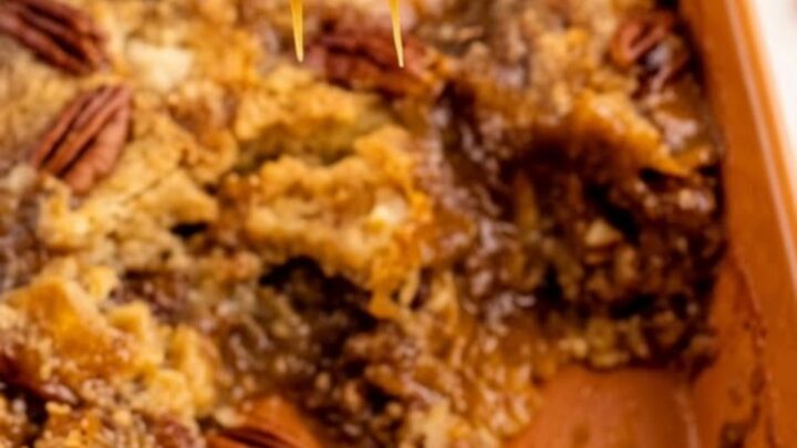 A close-up of a warm pecan pie dump cake in an orange ceramic dish with a spoon scooping out a gooey portion.