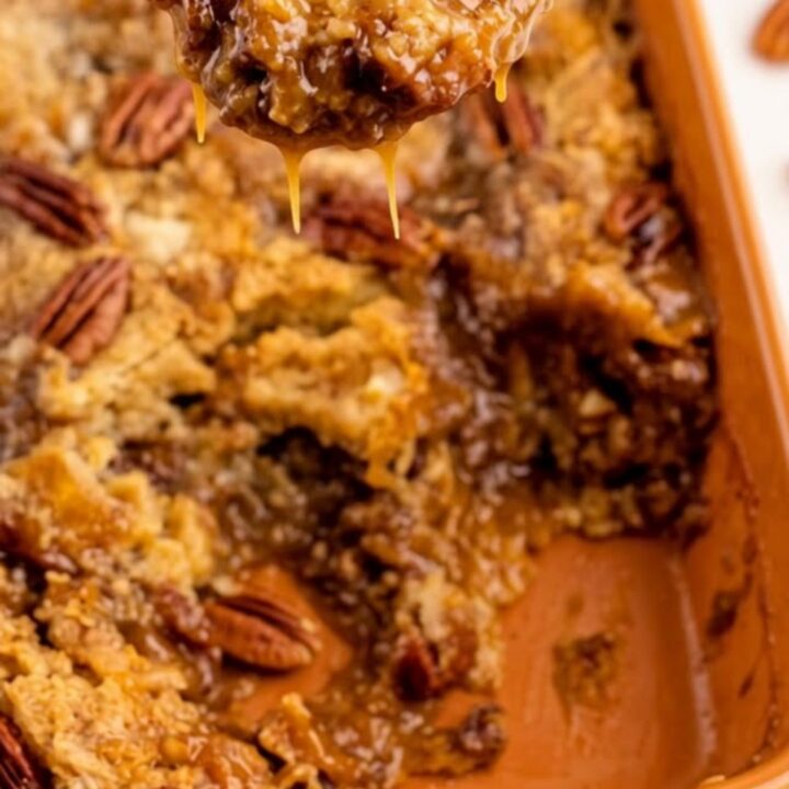 A close-up of a warm pecan pie dump cake in an orange ceramic dish with a spoon scooping out a gooey portion.