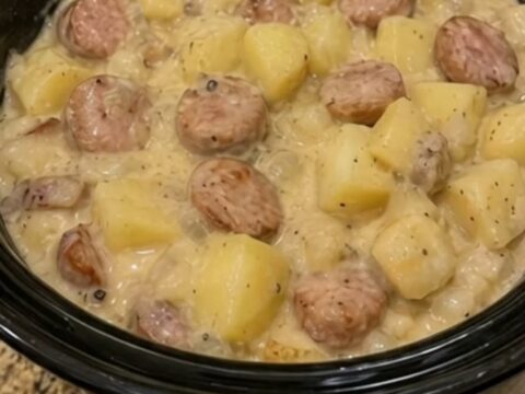 A close-up of a white bowl filled with creamy diced potatoes and sliced smoked sausage, steaming hot.