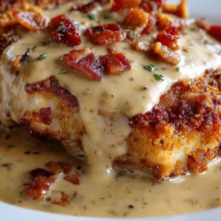 A golden-brown crispy pork chop on a white plate, generously topped with a thick, creamy bacon gravy and crispy bacon bits.