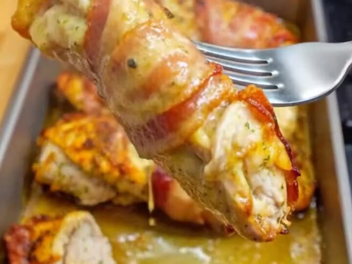 A steaming, bacon-wrapped chicken breast resting in a metal baking pan, showing crispy edges and a juicy interior.