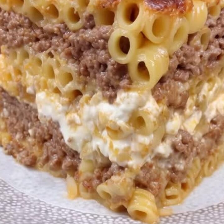 A tall, perfect square slice of layered Greek pastitsio on a white textured plate, steaming under warm golden light.