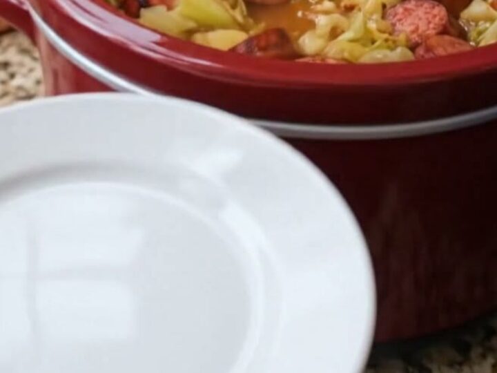 A close-up of a steaming portion of slow-cooked sausage, tender cabbage, and potatoes in a white bowl.