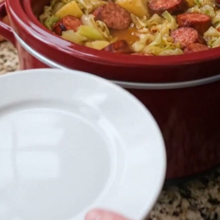 A close-up of a steaming portion of slow-cooked sausage, tender cabbage, and potatoes in a white bowl.