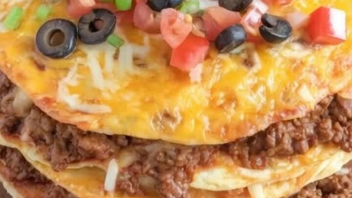 A towering homemade Mexican pizza on a round wooden board, topped with melted cheese, sour cream, diced tomatoes