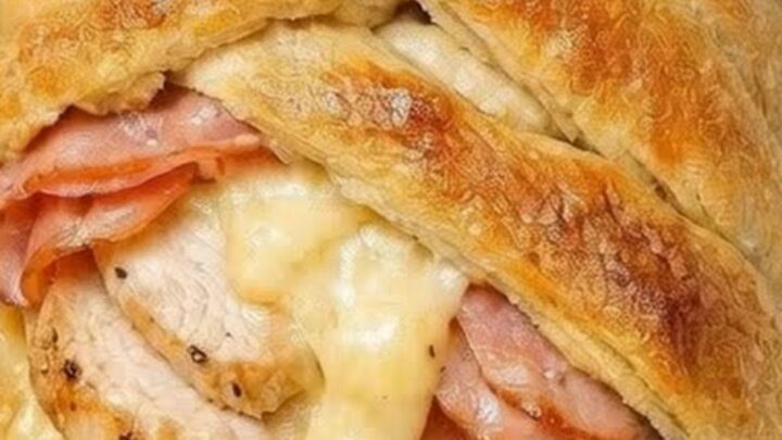A golden brown, braided crescent pastry filled with chicken, ham, and melted Swiss cheese on a baking sheet.