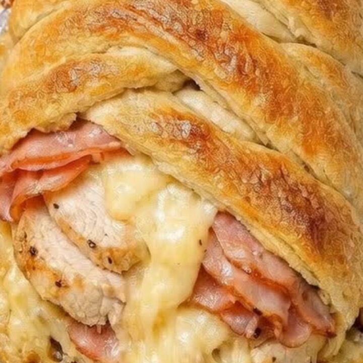 A golden brown, braided crescent pastry filled with chicken, ham, and melted Swiss cheese on a baking sheet.