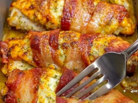 A golden, bacon-wrapped chicken breast resting in a metal baking pan with a crispy, savory crust.