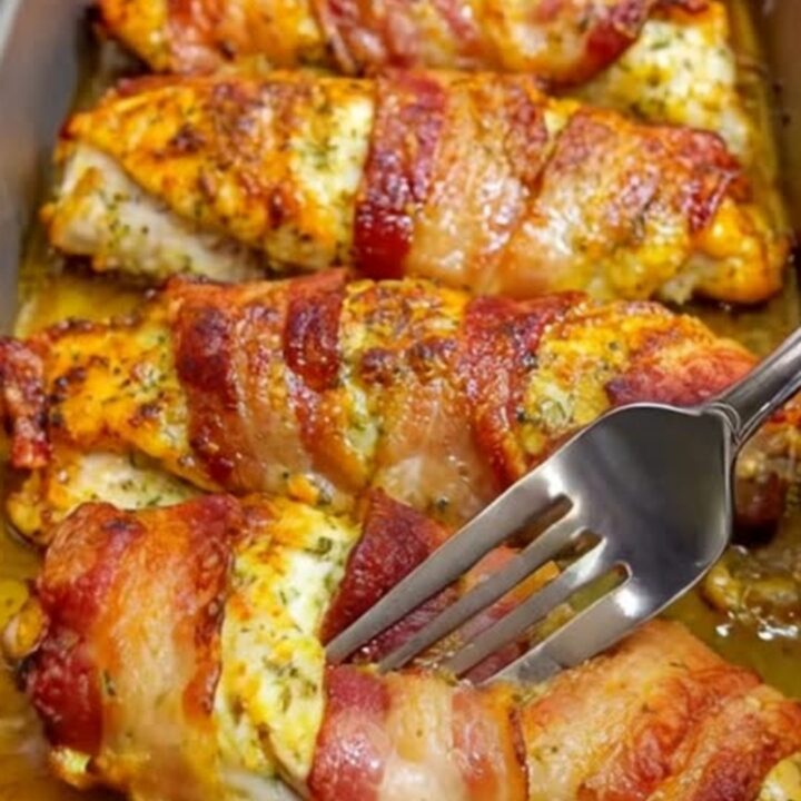 A golden, bacon-wrapped chicken breast resting in a metal baking pan with a crispy, savory crust.