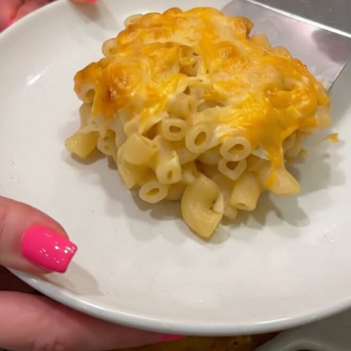 A close-up of a square of baked macaroni and cheese being lifted from a white baking dish with a metal spatula.
