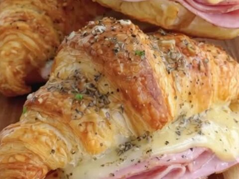 A close-up of a warm, golden, flaky ham and cheese croissant sandwich on a wooden board.